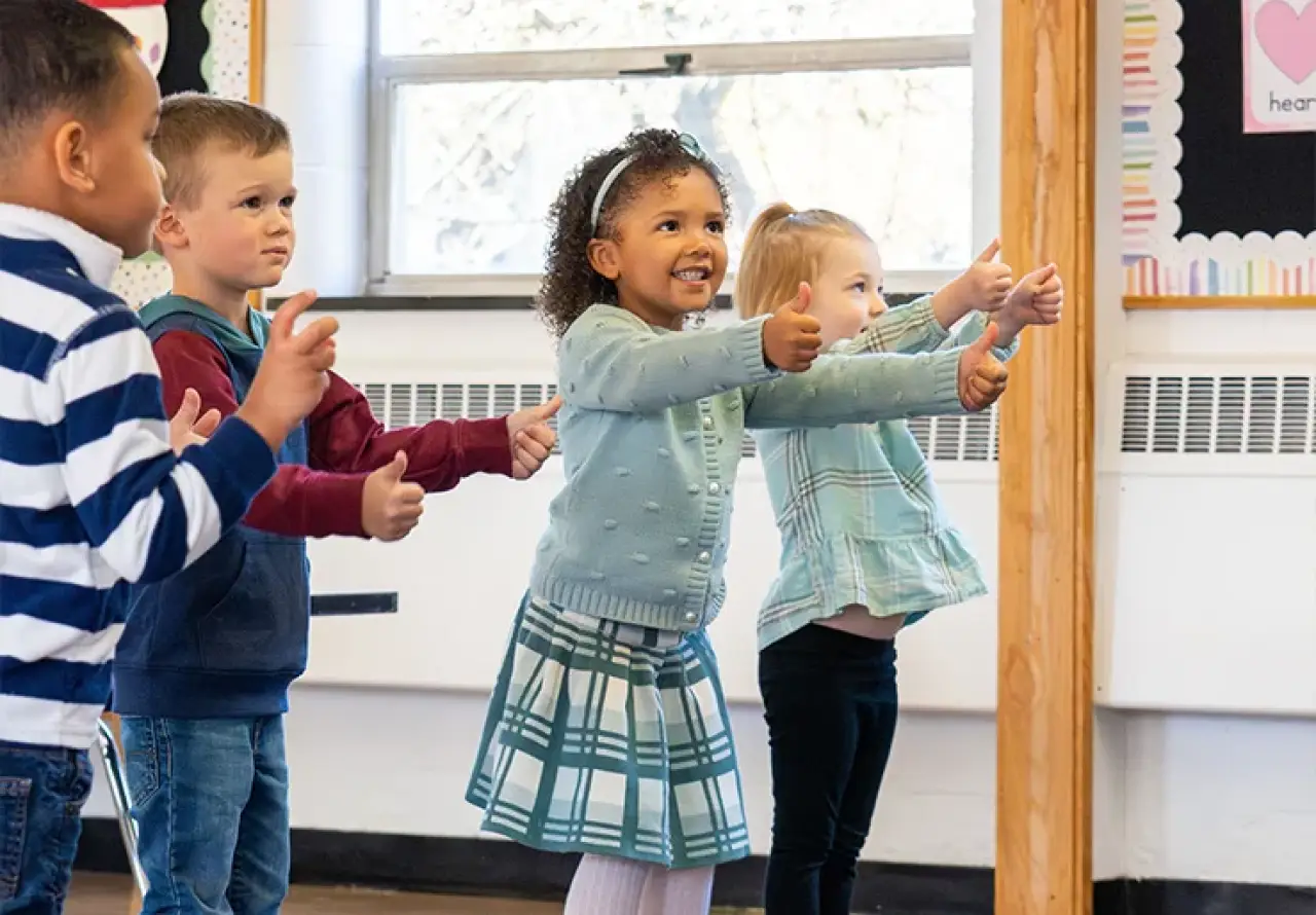 Children in a classroom standing in a row, giving thumbs-up gestures.