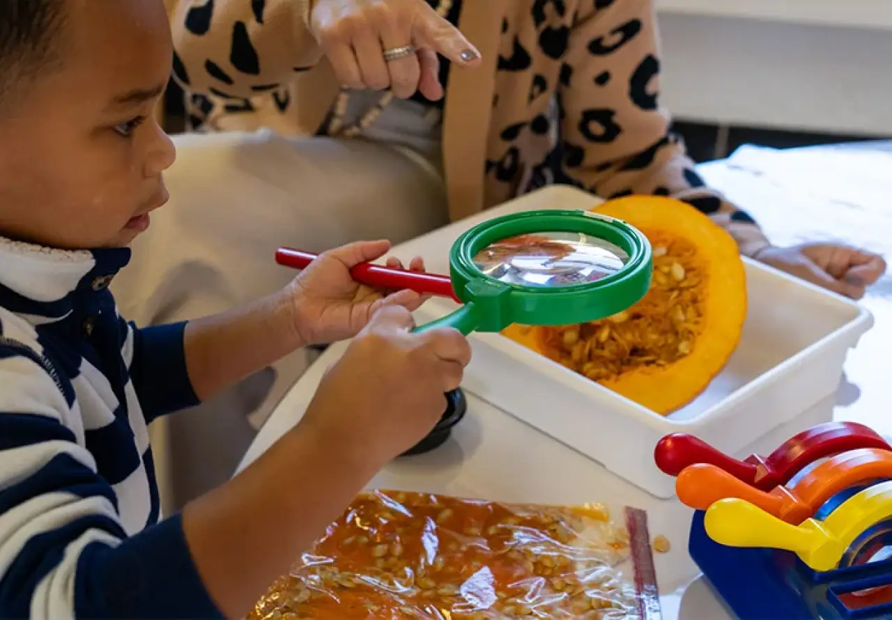 Child using a magnifying glass to explore pumpkin seeds on a table.