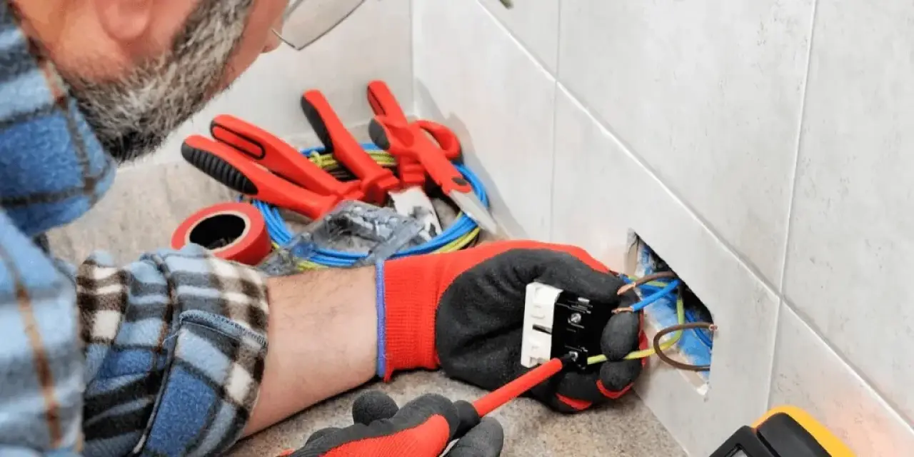Person fixing electrical wires in a wall socket with tools and gloves on a work surface nearby.