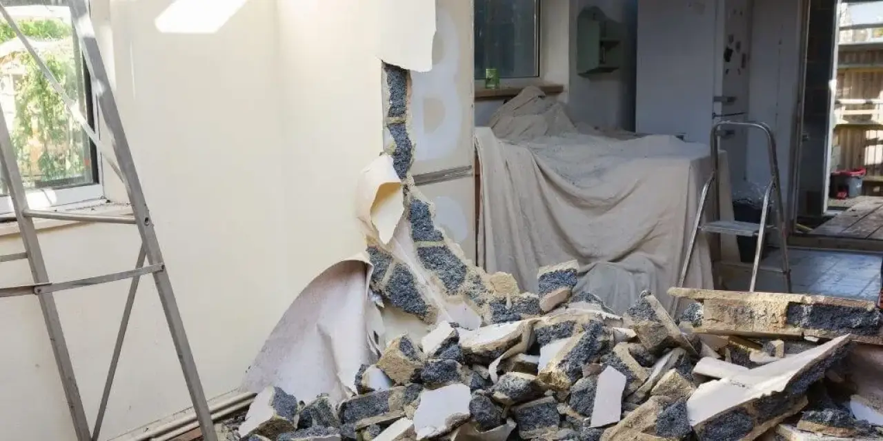 A wall is broken with debris on the floor; a covered sofa and ladder are in the background.