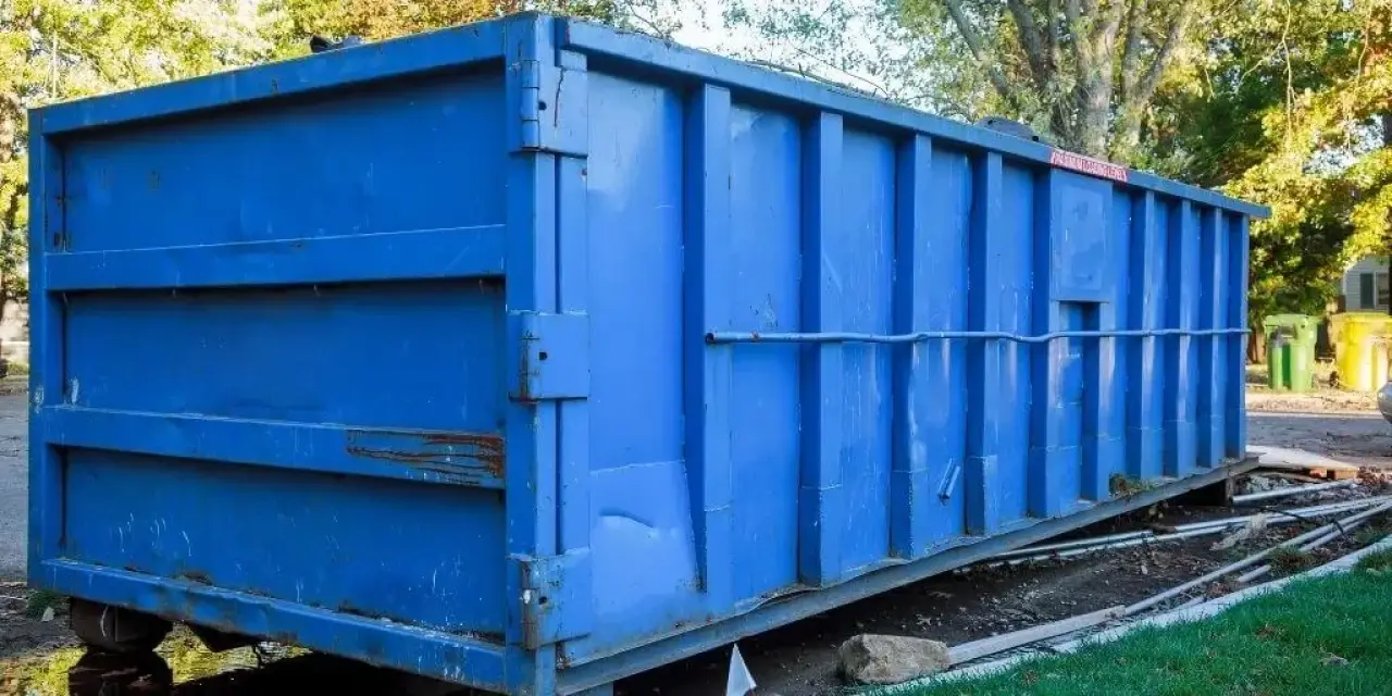 Large blue metal dumpster sits outdoors on a patch of dirt and grass near some trees.