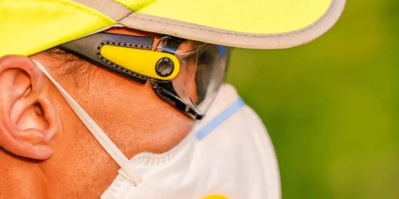 Person wearing a yellow cap, sunglasses, and a white face mask, shown in close-up profile.