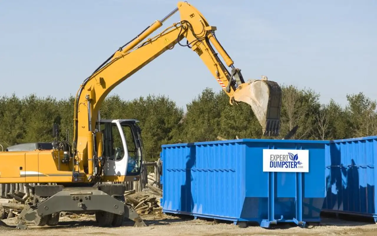 an excavator loads debris into a large blue dumpster at a construction site