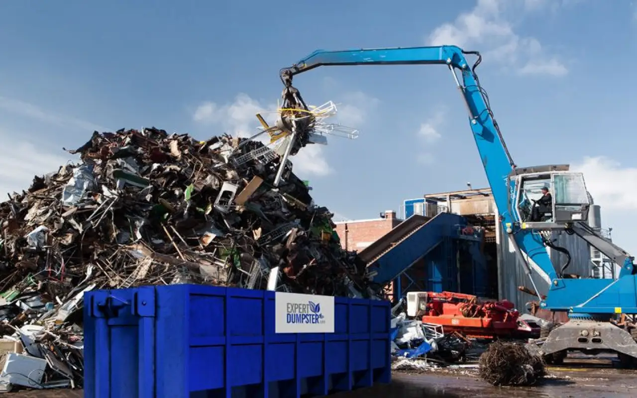 a blue excavator moves metal scrap into a large dumpster at a junkyard