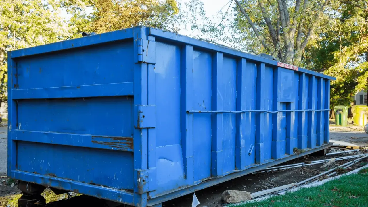 Large blue metal dumpster sits outdoors near grass and trees on a sunny day.