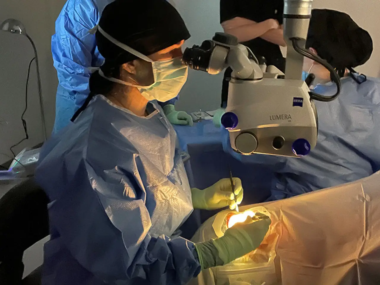 Surgeon in scrubs performing eye surgery using a microscope in an operating room.