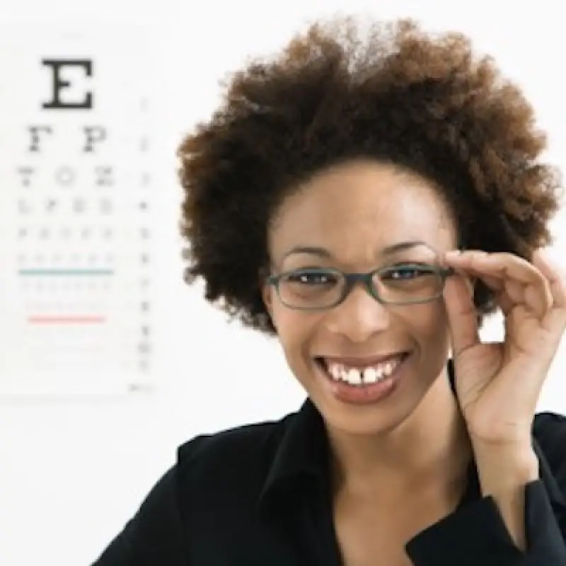 Smiling woman wearing glasses, standing in front of an eye chart at an optometrists office.