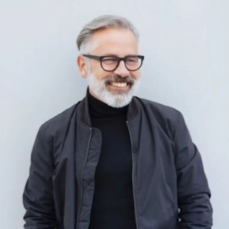 Smiling man with gray hair and beard wearing glasses, a black turtleneck, and a jacket.