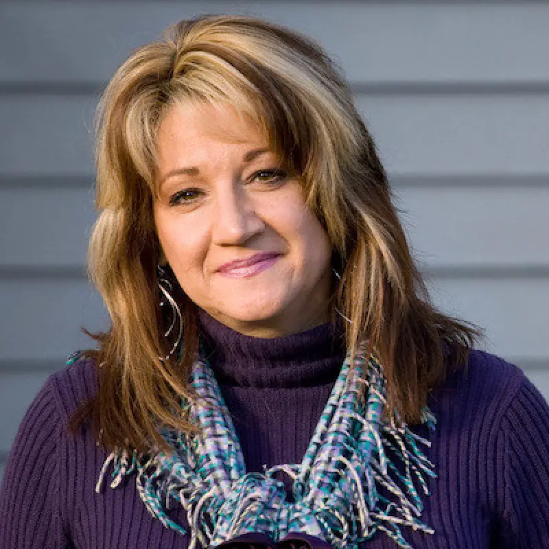 Woman with light brown hair in a purple sweater and scarf, smiling in front of a gray wall.