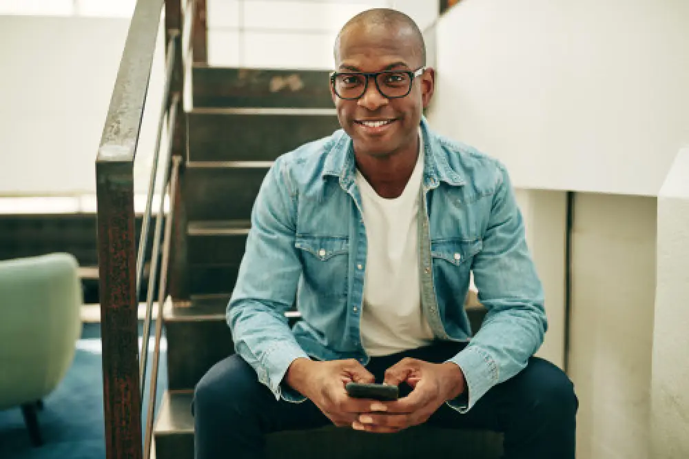 A black man wearing a jean jacket and glasses sitting on a staircase and smiling.