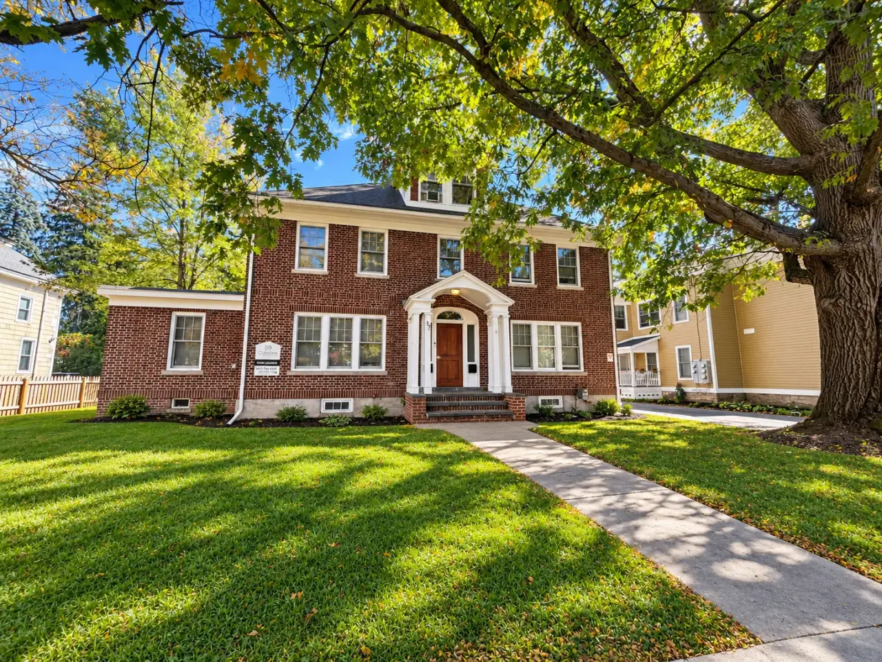 A two-story brick house with a brown front door, surrounded by a lawn and mature trees.
