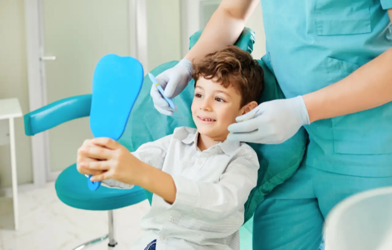 Smiling boy holding a mirror at the dentists office with a dental professional beside him.