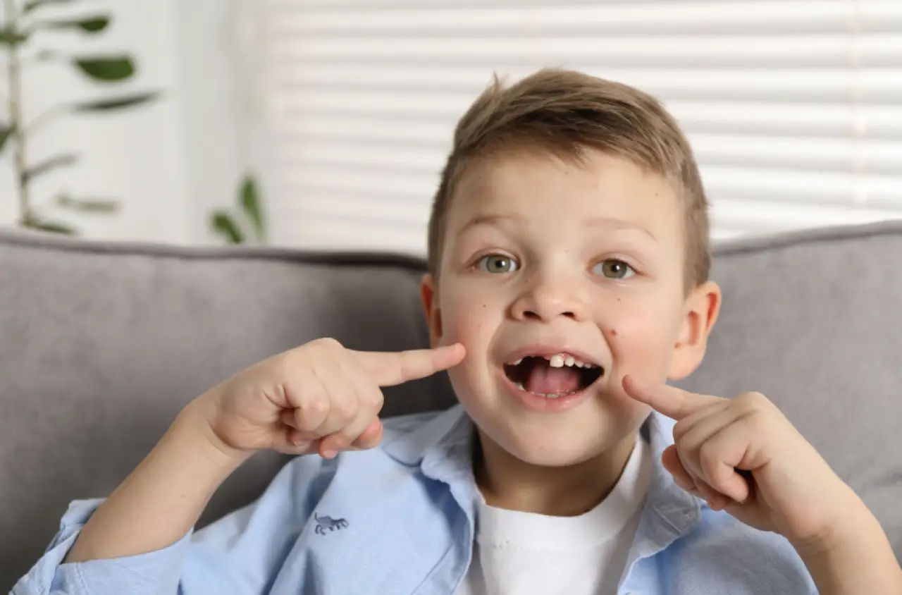 Smiling boy pointing to his missing front teeth while sitting on a gray couch indoors.