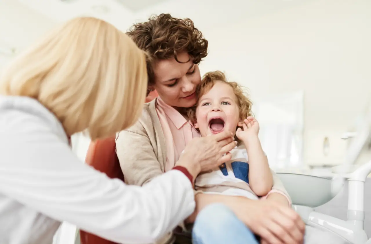 A smiling child sits on an adult’s lap as a dentist examines the child’s open mouth.