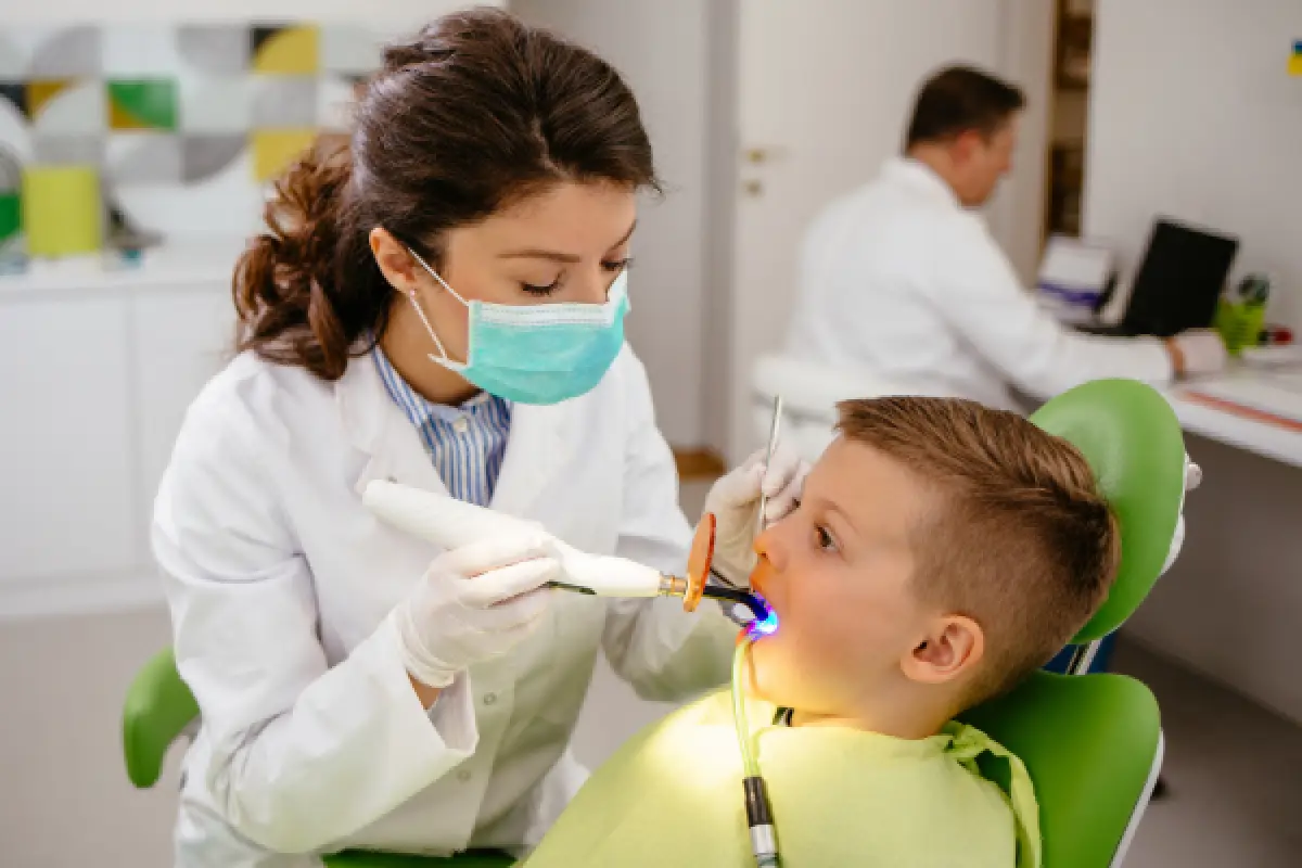 Dentist in a mask examines a young boy’s teeth with dental tools in a clinic.