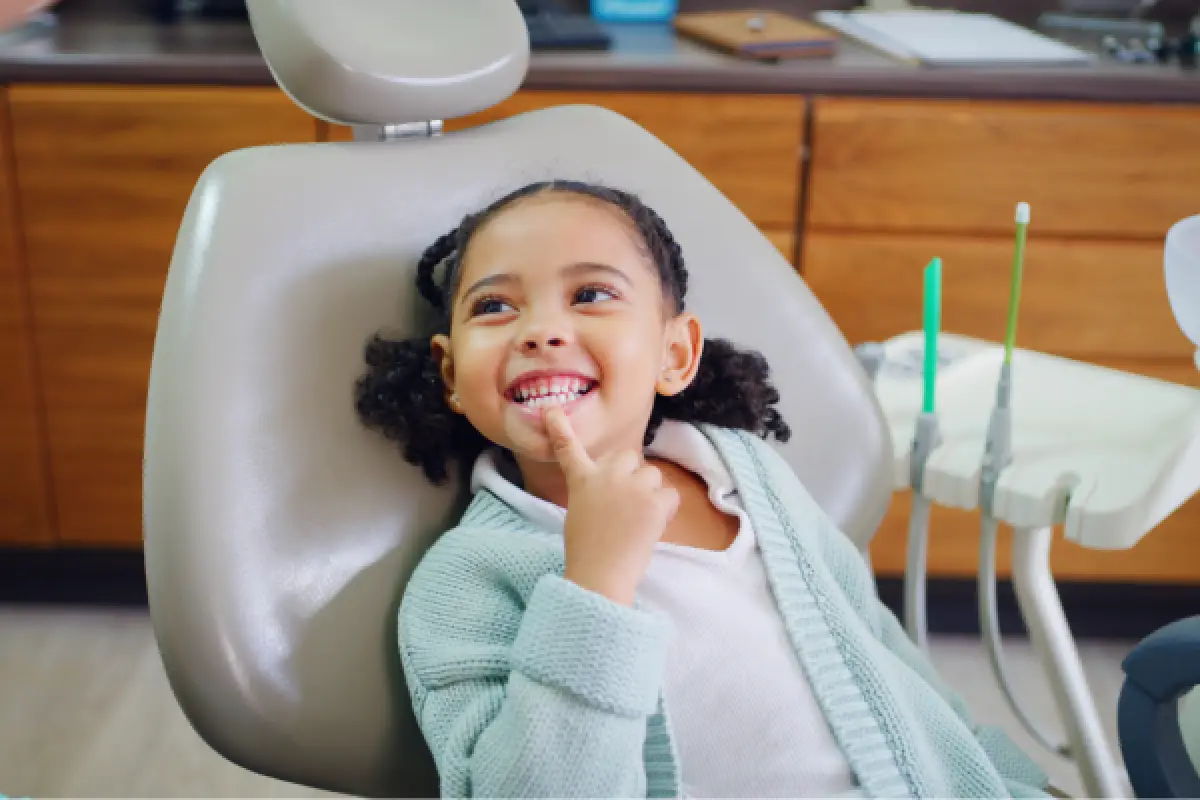 Smiling young girl sits in a dentist chair, pointing to her teeth in a dental clinic.