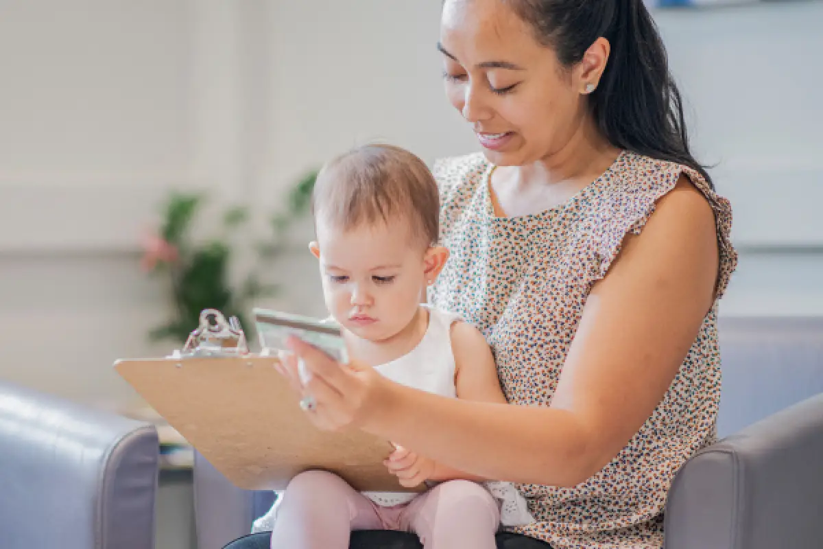 Woman and baby sit together, looking at a clipboard and phone in a waiting room.