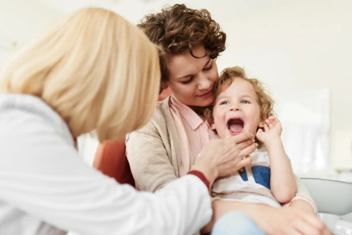 A child sits on a woman’s lap while a doctor checks the child’s mouth in a clinic.