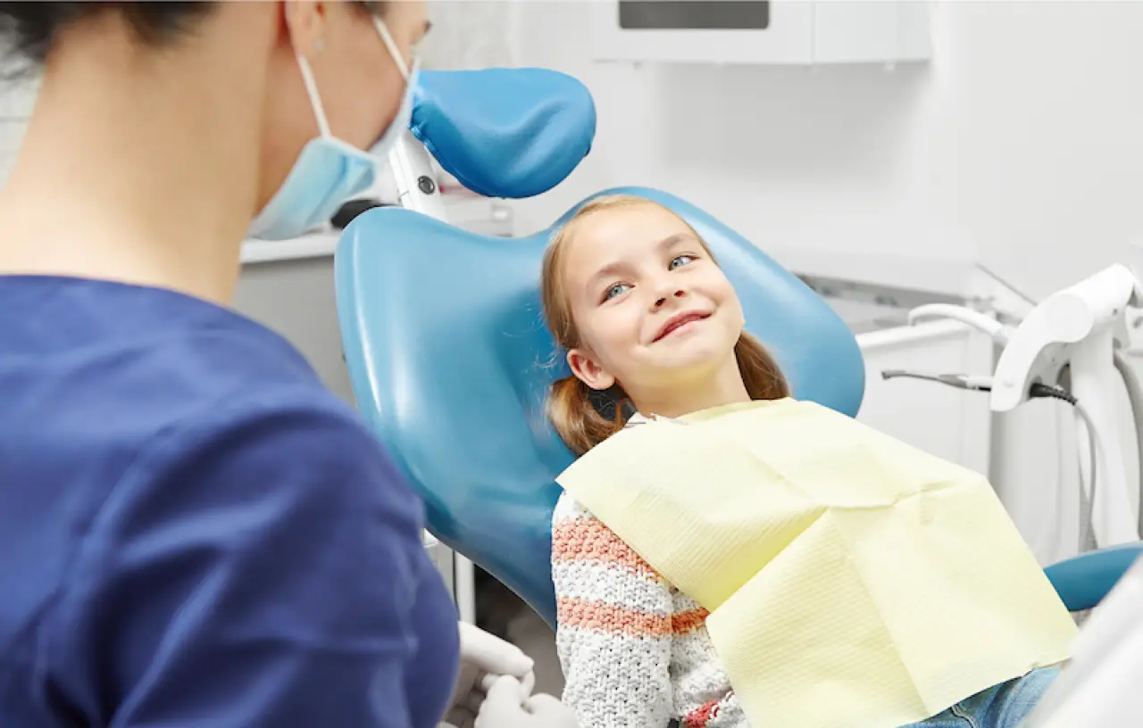 A young girl smiles while sitting in a dentists chair, with a dental professional nearby.