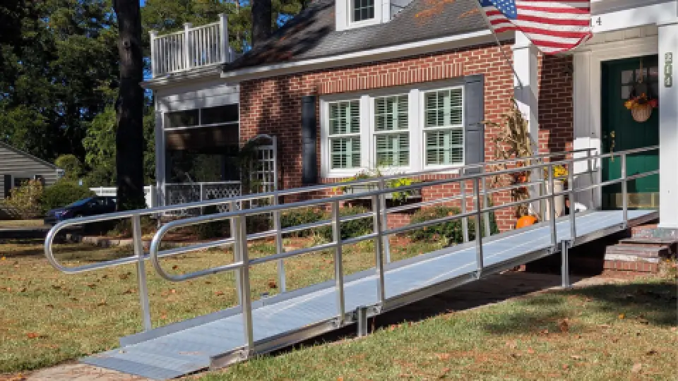 A metal wheelchair ramp leads to the front door of a brick house with an American flag.