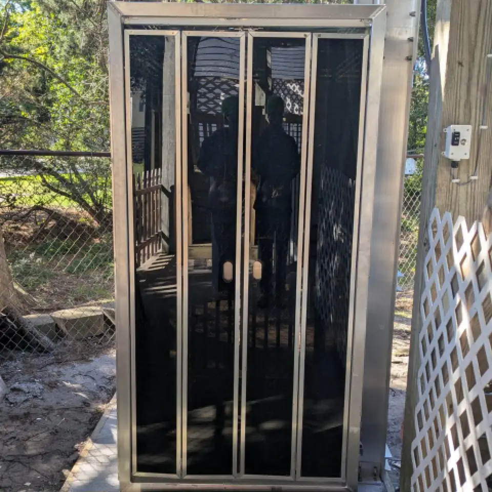 A metal outdoor shower enclosure with black glass doors, reflecting trees and a wooden deck.