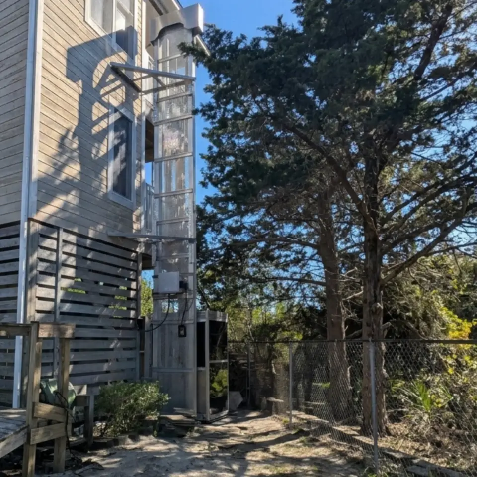A glass outdoor elevator attached to a wooden house beside a tall tree and chain-link fence.