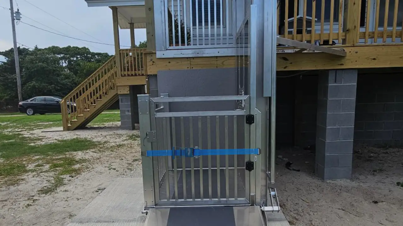 Outdoor wheelchair lift with metal gate and ramp, installed beside a raised wooden house.