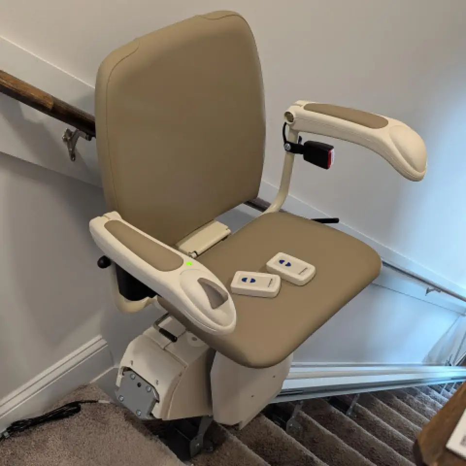 A beige stairlift with two remote controls is installed on a staircase with a handrail.