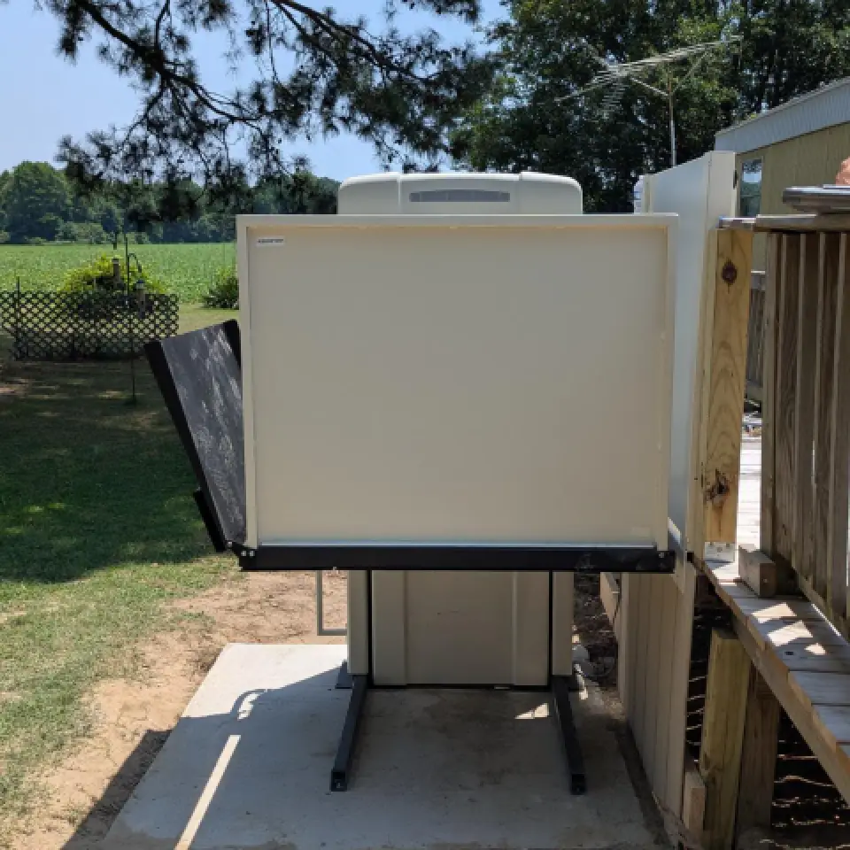Vertical wheelchair lift outside, next to a wooden ramp and mobile home, on a sunny day.