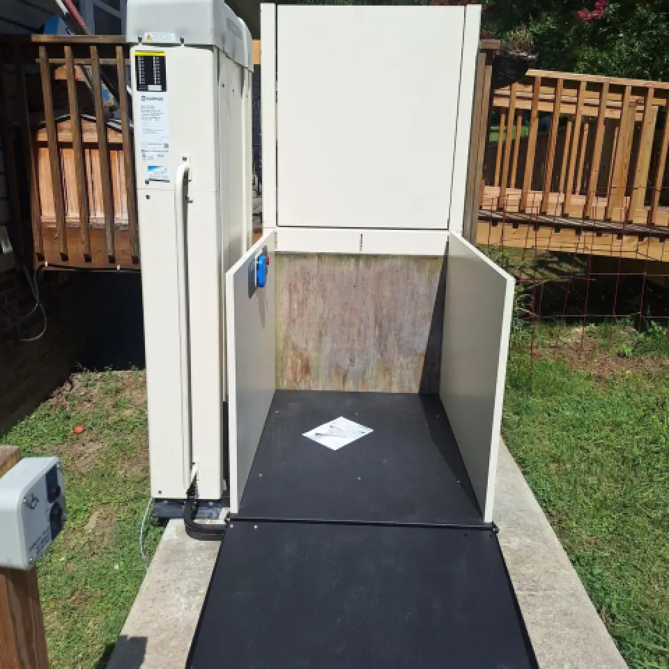 Vertical platform lift with a black ramp, outdoors near a wooden deck, on a concrete base.