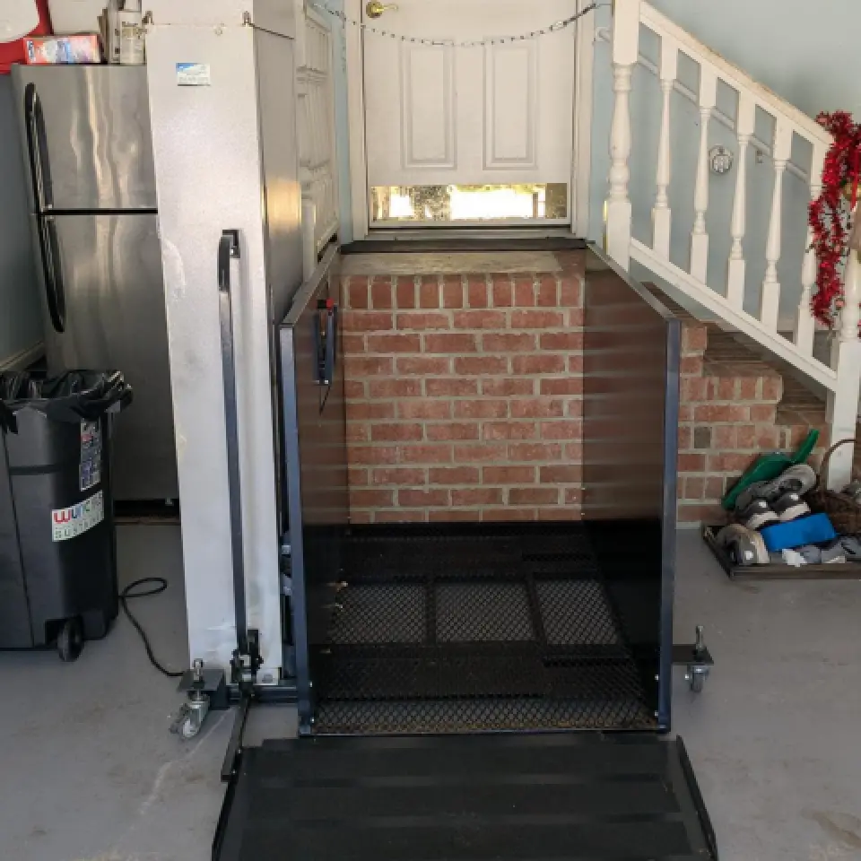 A wheelchair lift leads up to a door next to a brick wall and a staircase with shoes nearby.