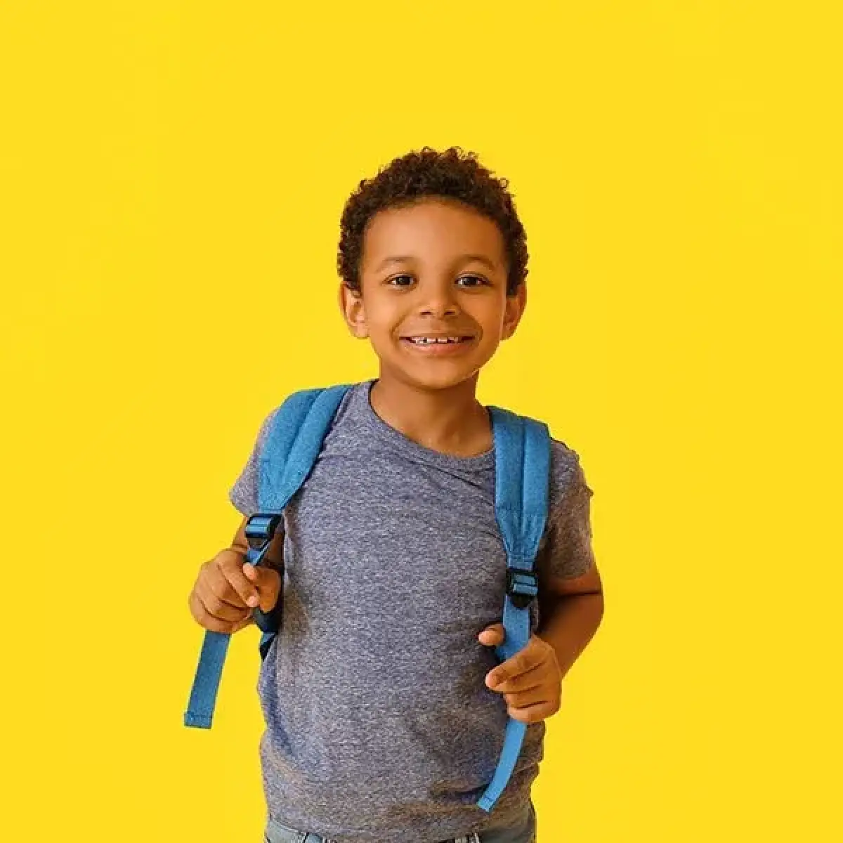 Smiling young boy with a blue backpack stands against a bright yellow background.