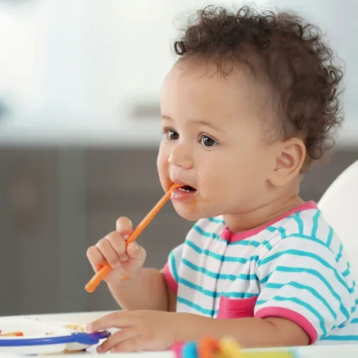 A toddler in striped pajamas holds an orange spoon and eats at a table.