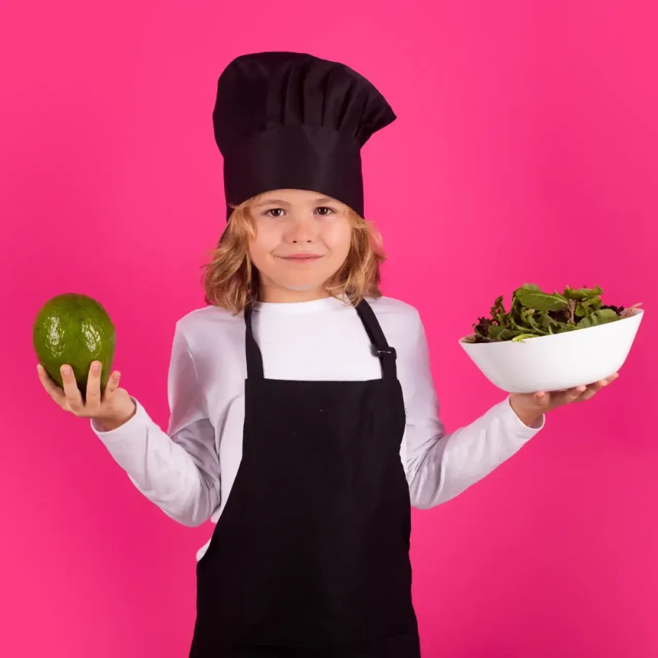 Child in chef hat and apron holding an avocado and a bowl of salad, standing on pink background.