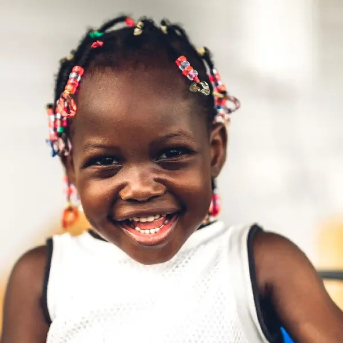 Smiling young girl with beaded braids, wearing a white sleeveless top, against a blurred background.