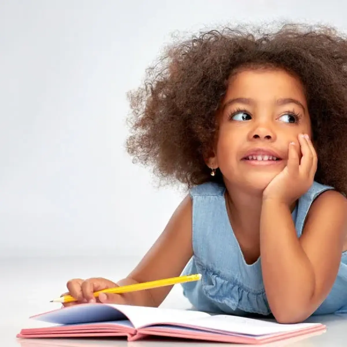 Young girl smiling and daydreaming while holding a pencil and sitting with an open notebook.