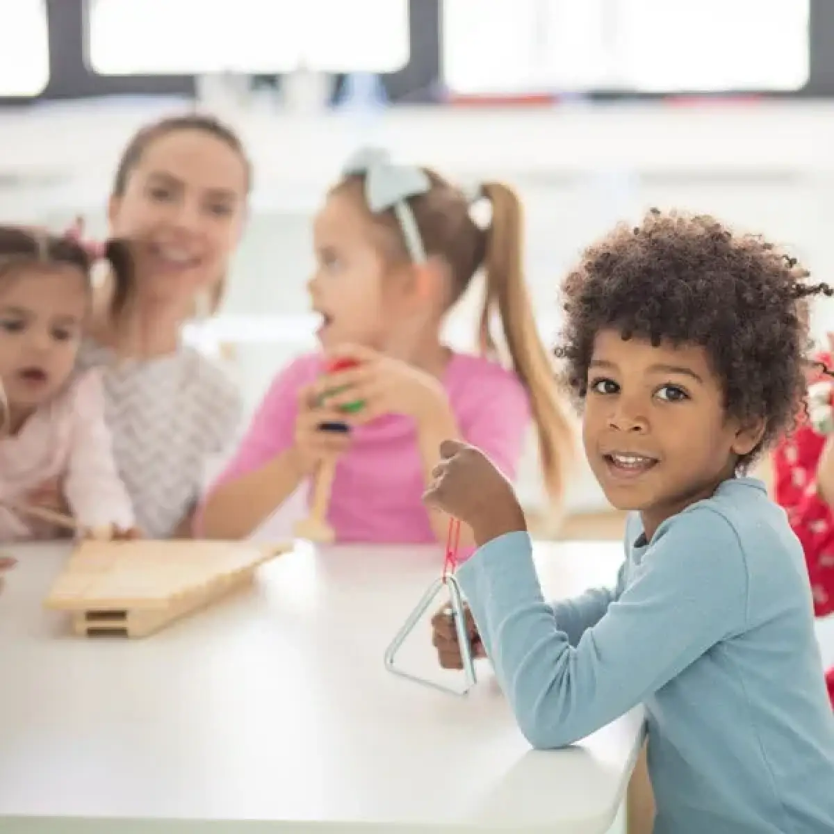 Smiling boy holding a triangle instrument at a table with other children and an adult.