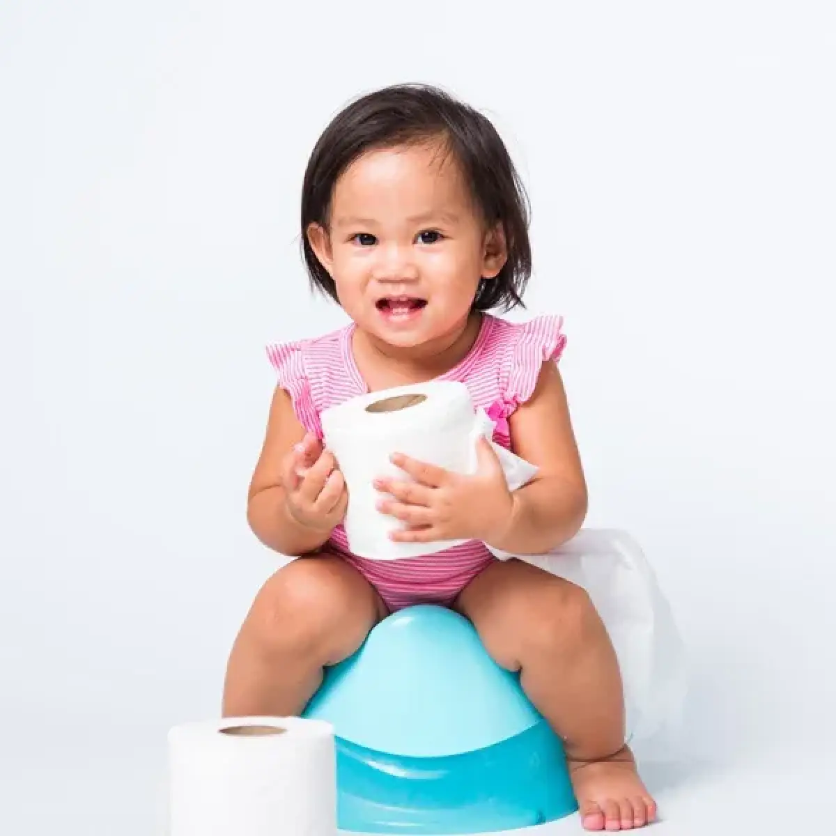 Smiling toddler in pink outfit sits on a potty, holding toilet paper rolls.