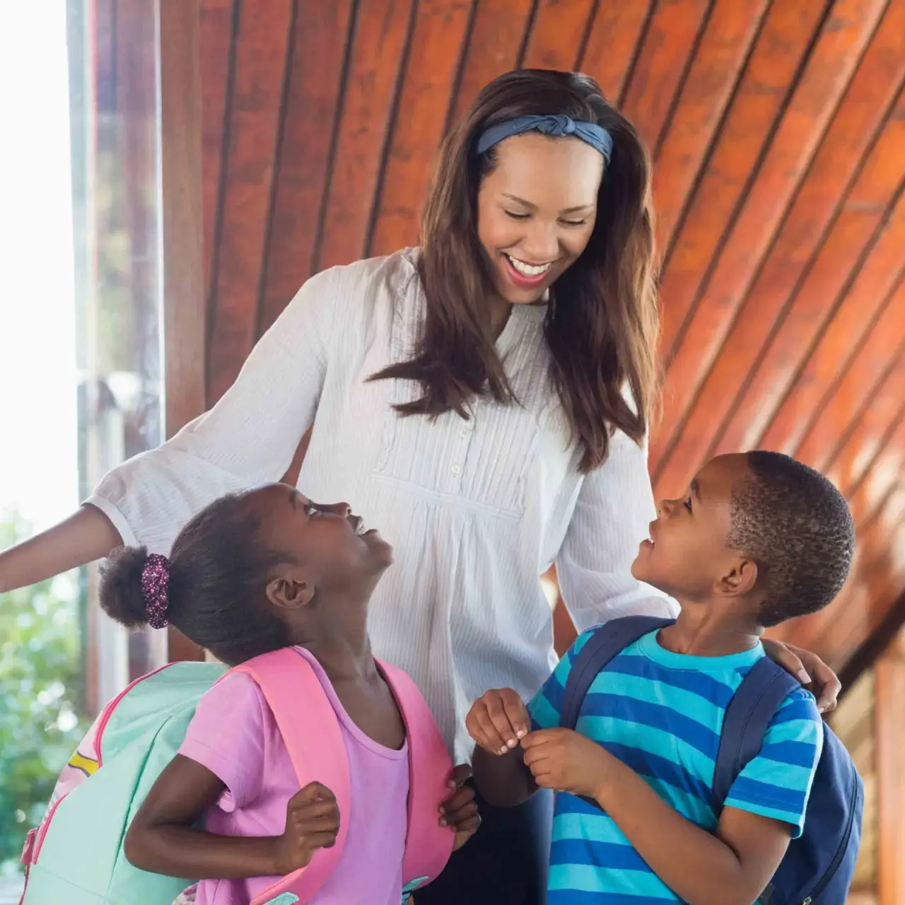 Smiling woman with two young children wearing backpacks, standing indoors and talking together.