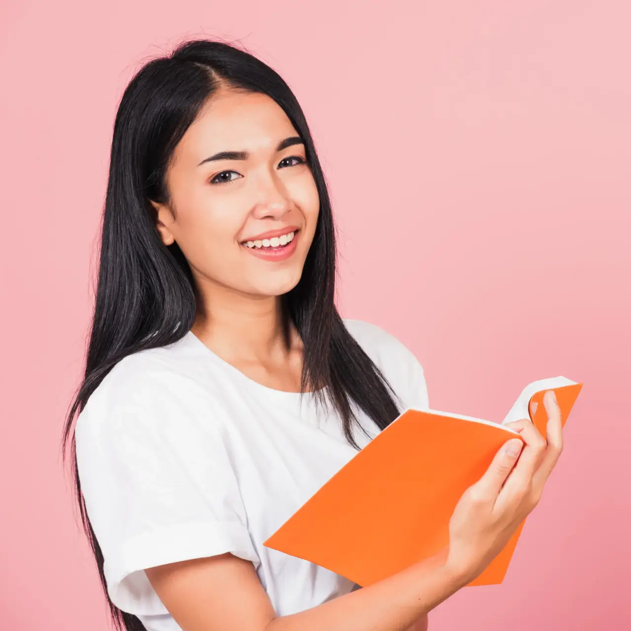 Woman with long black hair smiling and holding an open orange book against a pink background.
