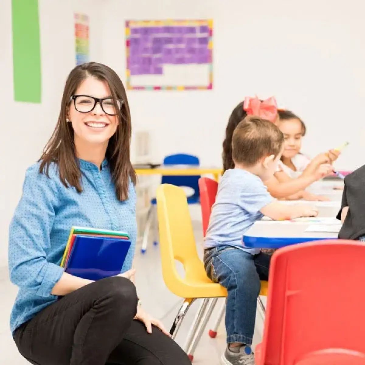 Smiling teacher with books kneels by young children sitting and drawing at a classroom table.