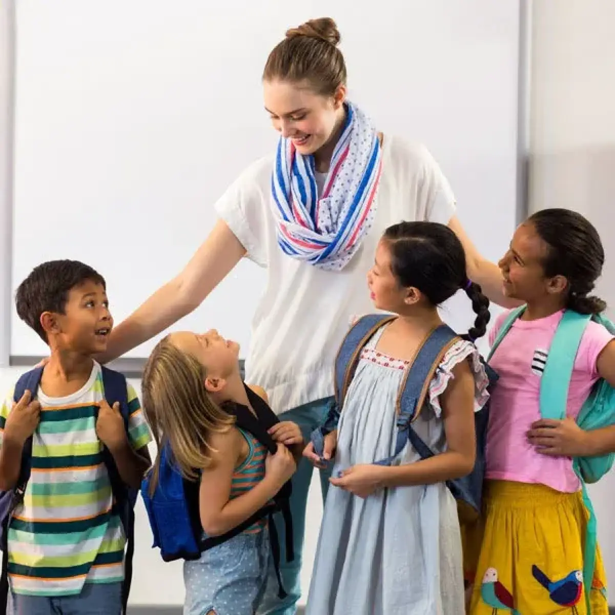 A teacher smiles with four young students wearing backpacks in a classroom.