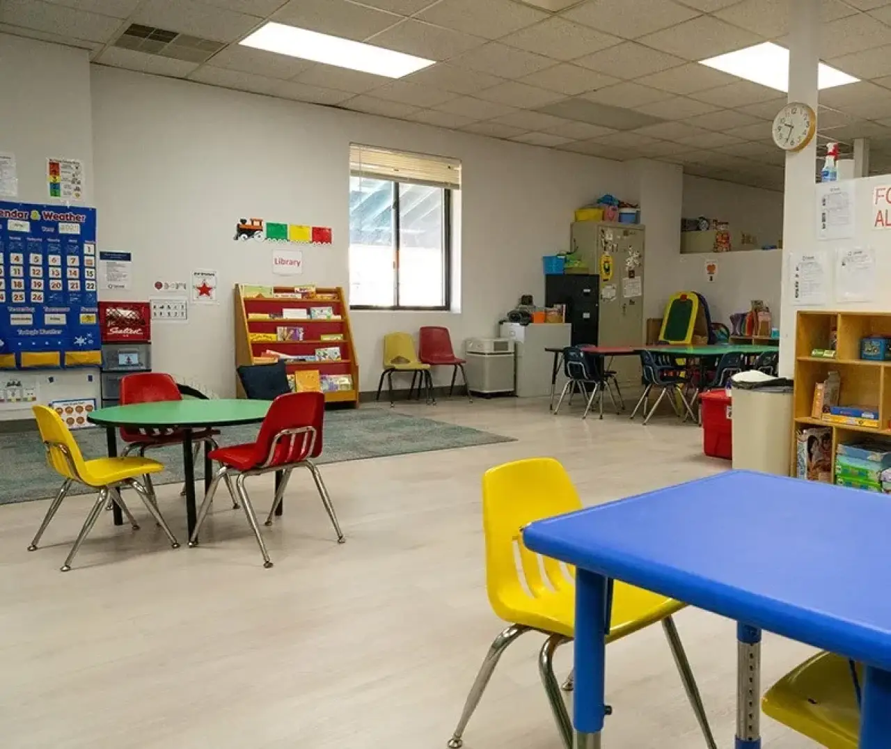 A colorful, empty classroom with small tables, chairs, bookshelves, and educational posters.