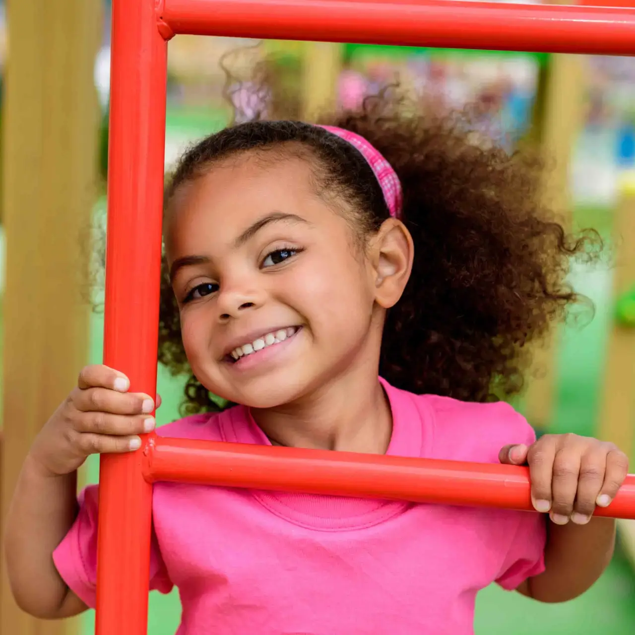 Smiling young girl in a pink shirt holding onto red bars at a playground.