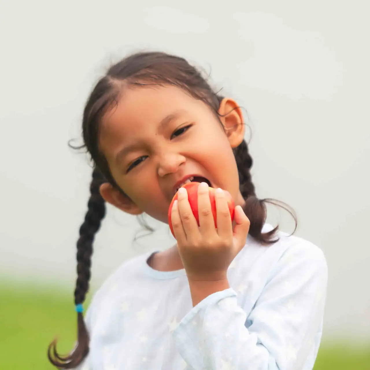 Young girl with braided hair taking a bite from a red apple, outdoors in soft light.