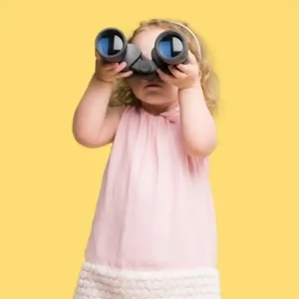 Young child in a pink dress looking through binoculars against a yellow background.