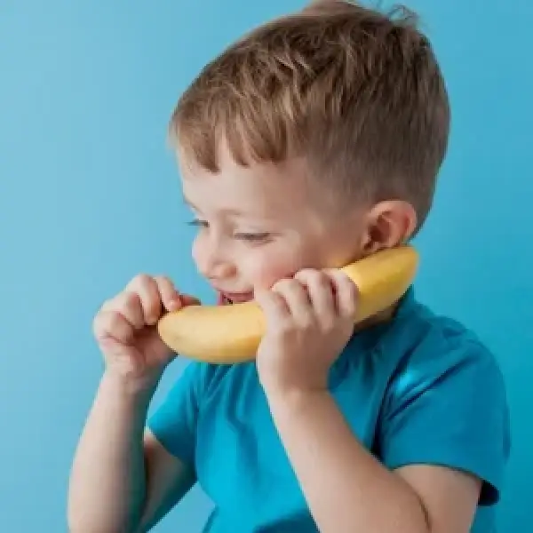 Smiling boy in a blue shirt holding a banana to his face like a phone against a blue background.