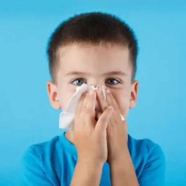 Young boy in a blue shirt blowing his nose with a tissue against a blue background.