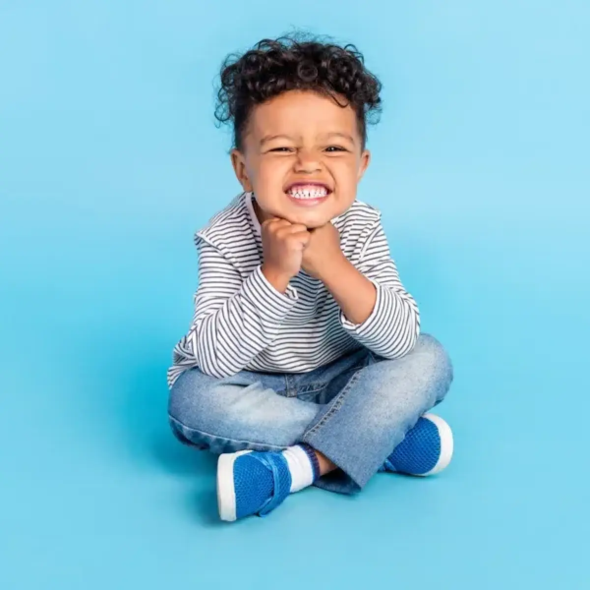 Smiling child in striped shirt and blue shoes sits cross-legged on a blue background.