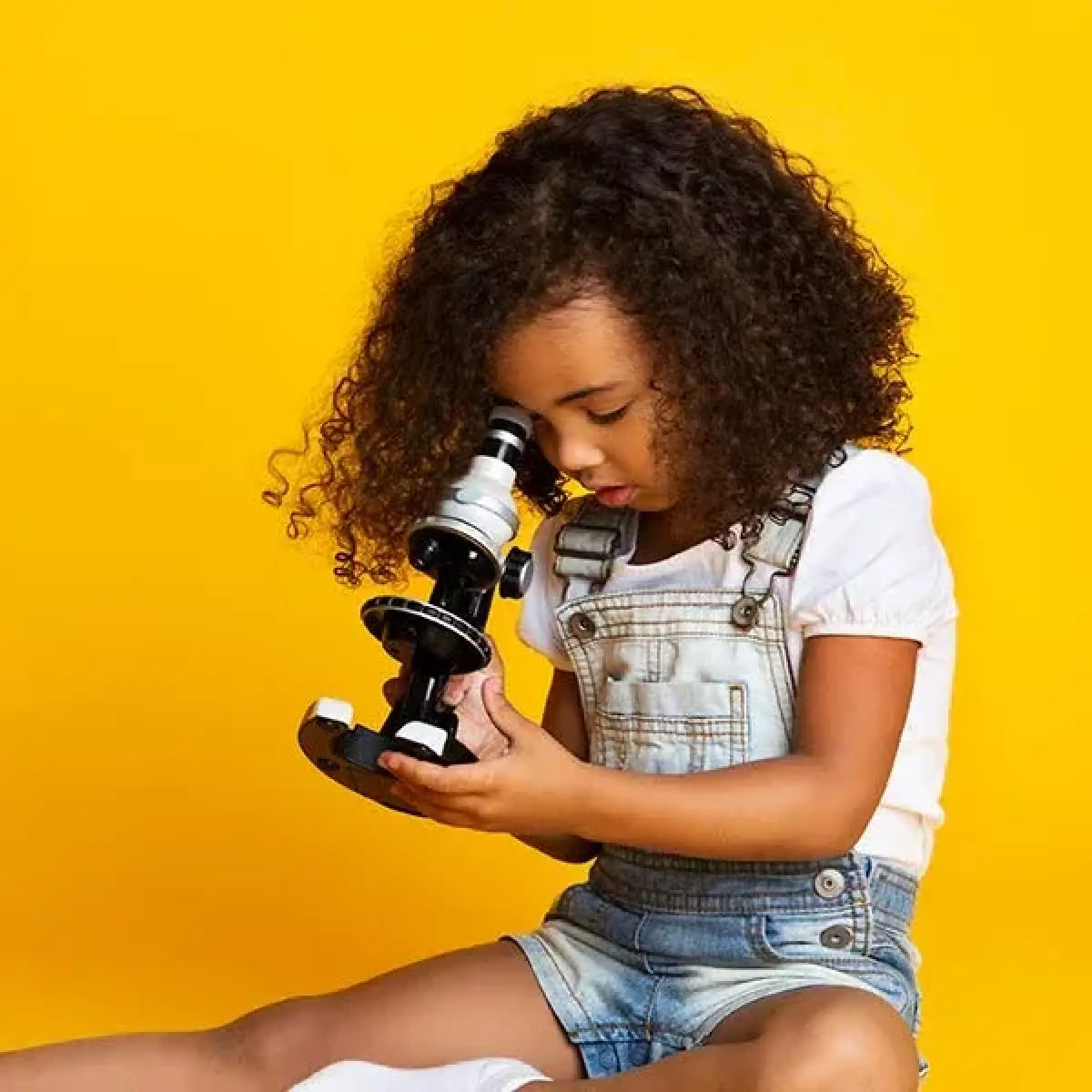 Young girl with curly hair looks through a microscope against a bright yellow background.